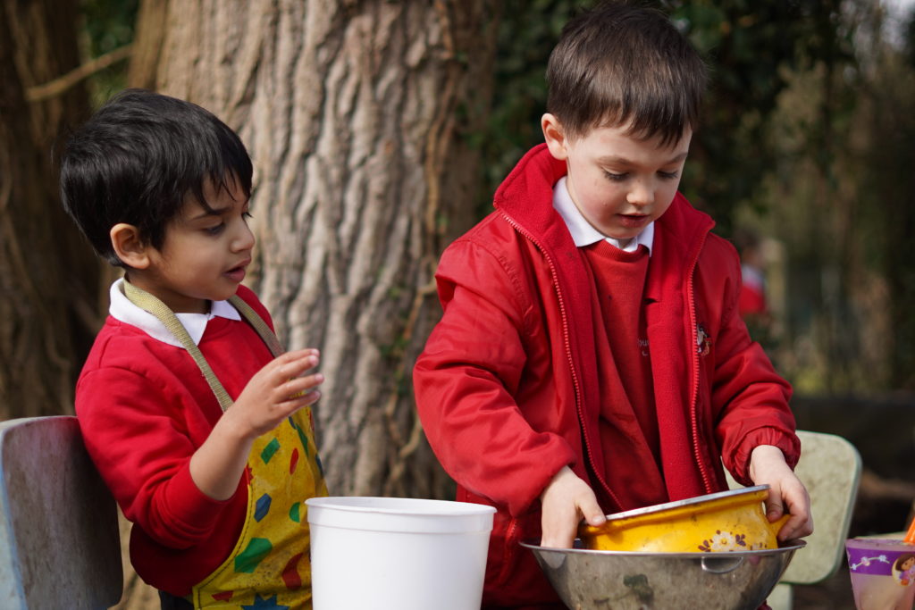 Two young boys in school uniform wearing yellow aprons while playing with pots outside on the playground