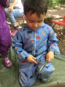 A young boy from the Nursery can be seen sitting in the Forest School area, using a knife to create stick men with tree branches.