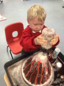 A young Nursery boy is shown pouring chemicals over a cone-shaped model to create the effect of a Volcano.