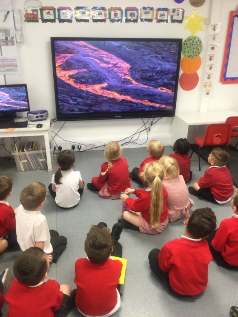 A class of Nursery pupils are shown sitting on the classroom floor and looking at the display board at the front of the room.