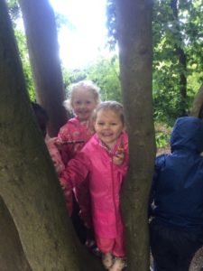 Two young girls from the Nursery can be seen huddled amongst the tree trunks, wearing their pink raincoats in the academy's Forest School area.