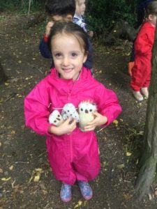 A young Nursery girl is shown wearing a pink raincoat and holding her soft toys in the Forest School area.