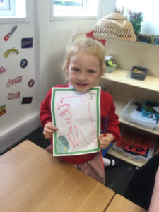 A young Nursery pupil is seen holding up a drawing of a Dinosaur that she has made.