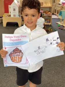 A young boy from Nursery is shown holding up some designs he has made of cakes as part of Enterprise Week.