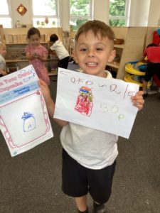 A young boy from Nursery is shown holding up some designs he has made of cakes as part of Enterprise Week.